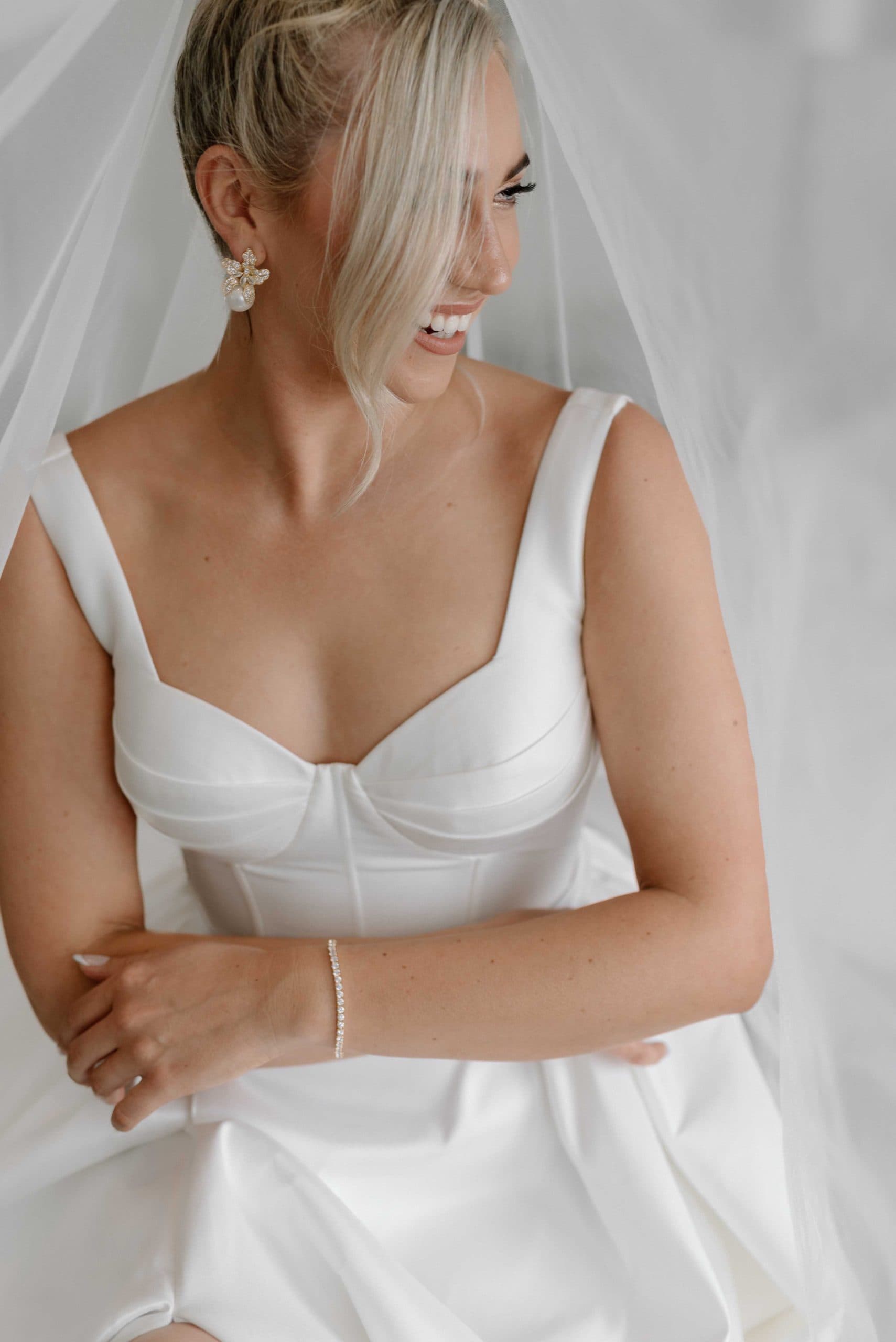 A smiling bride with short blonde hair wears a white corset gown and veil