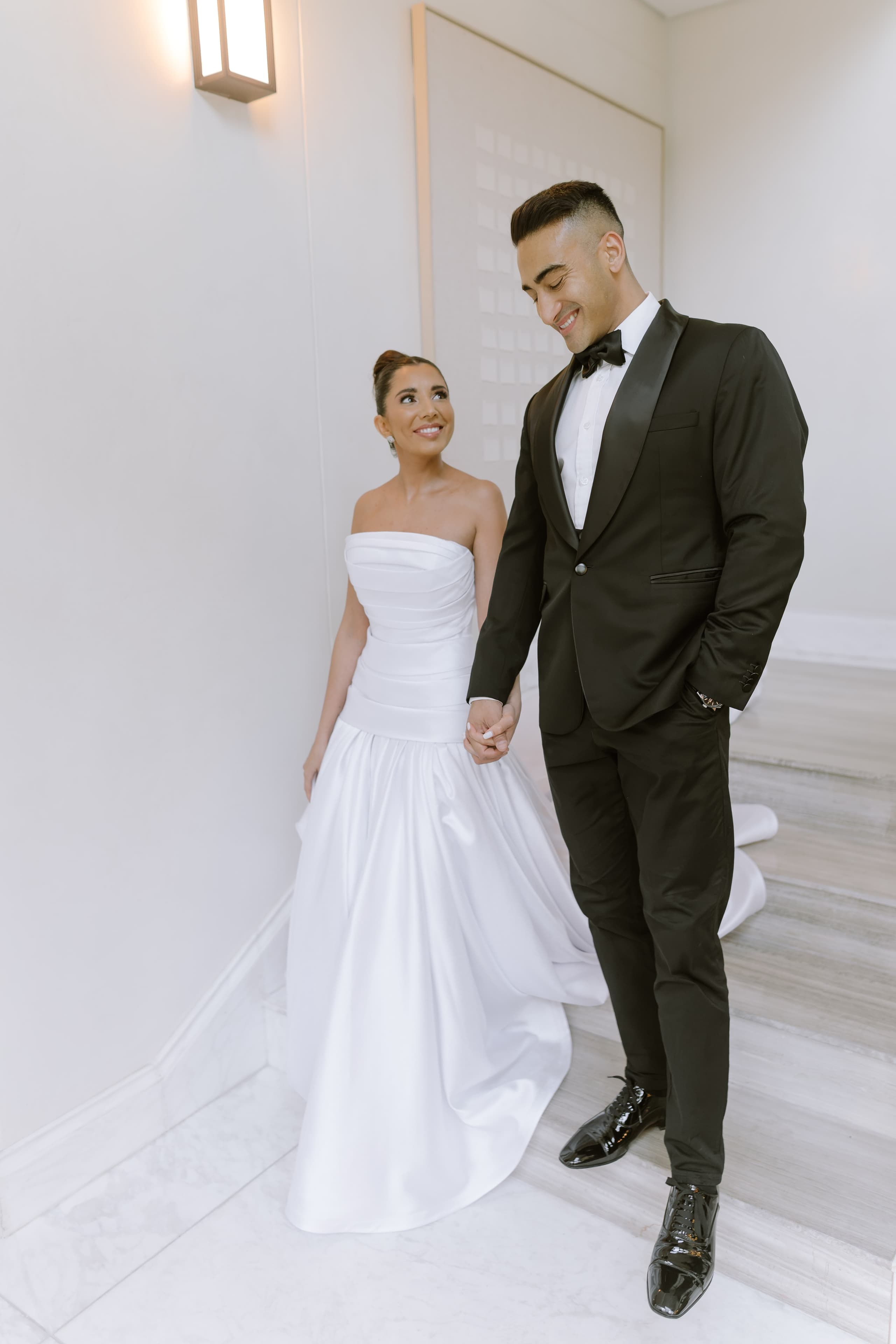 A smiling bride in a strapless gown holds hands with her groom in a tuxedo