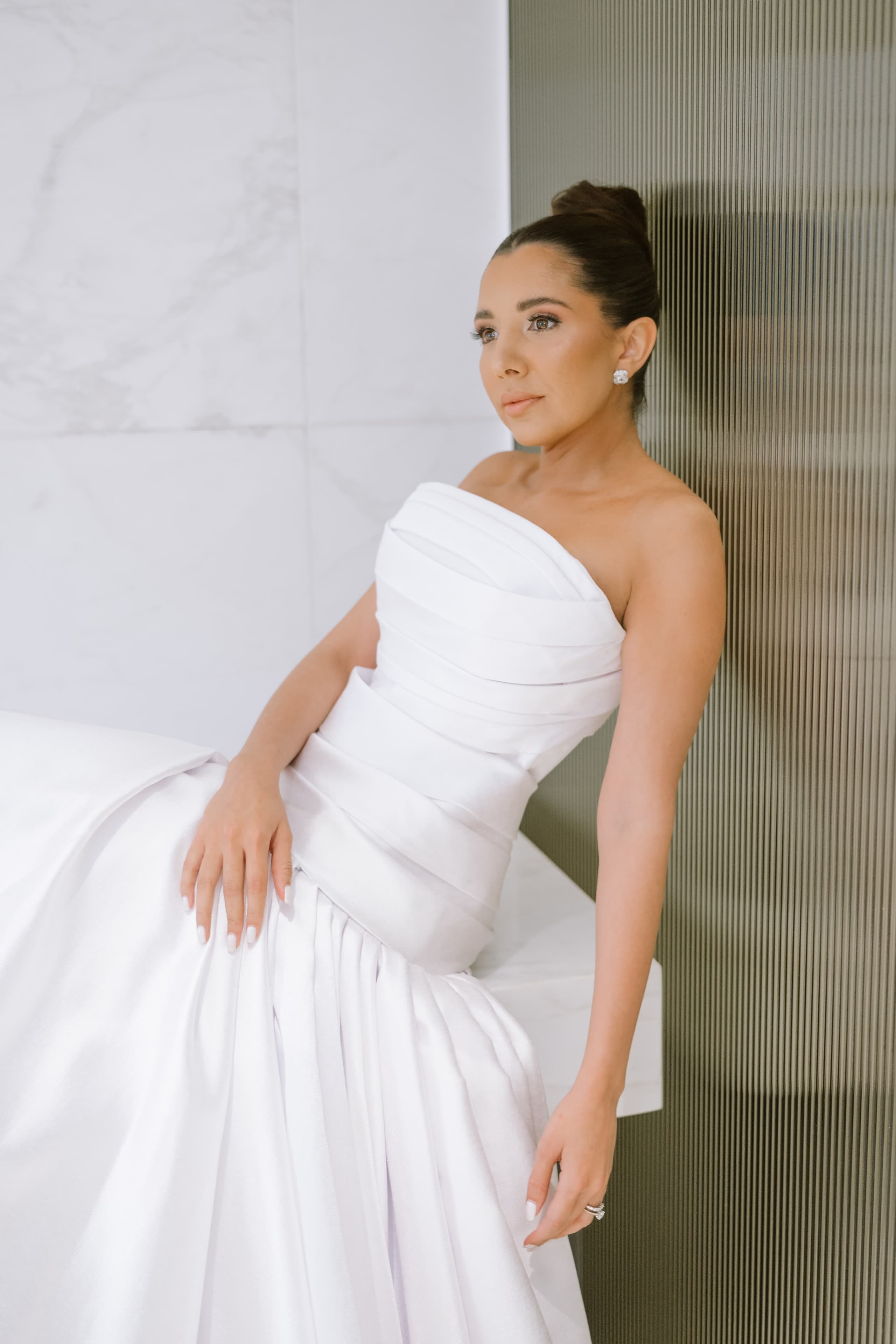 A bride in a pleated strapless white gown sits gracefully against a modern textured wall