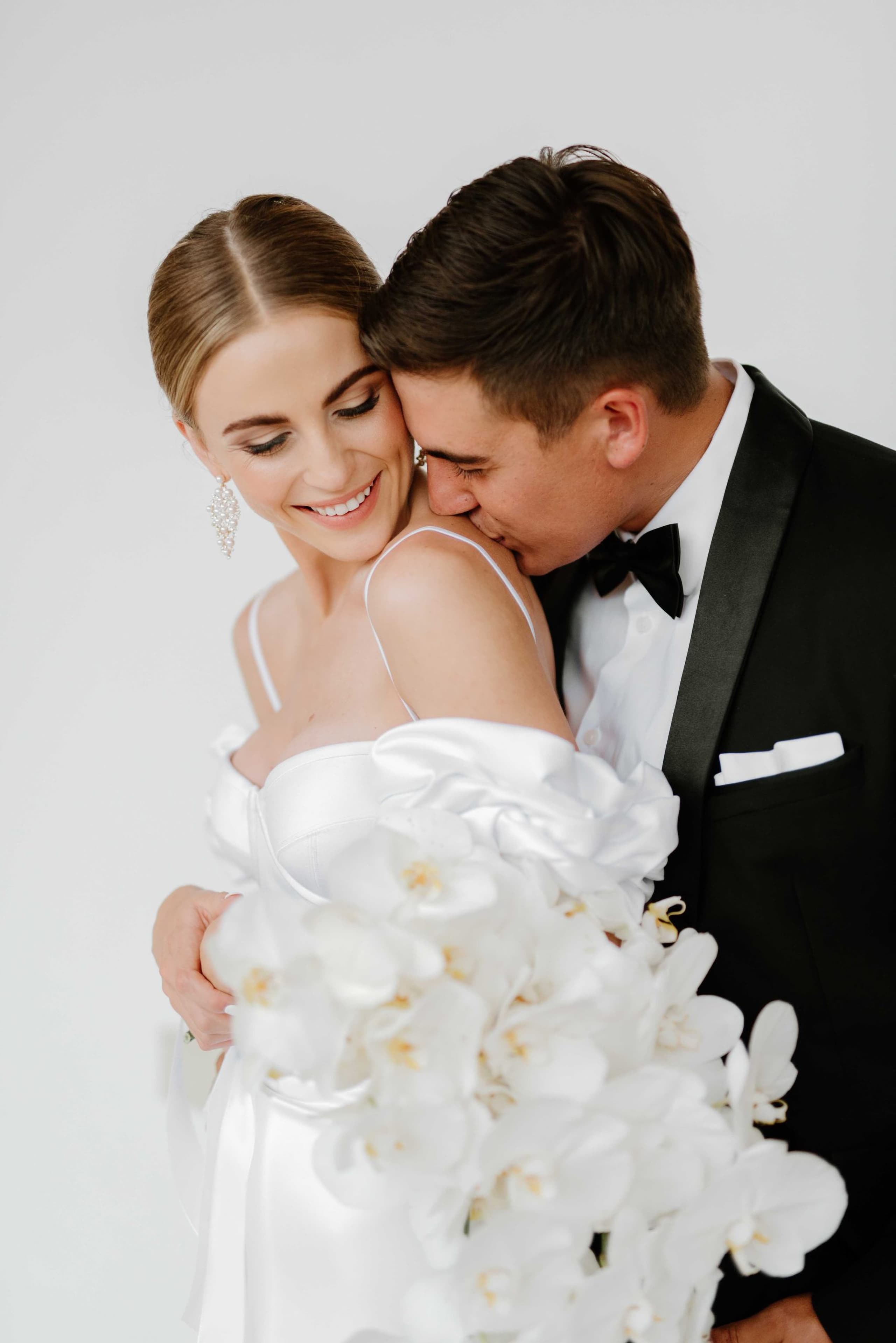 A groom kisses his bride on the shoulder while she holds a white orchid bouquet