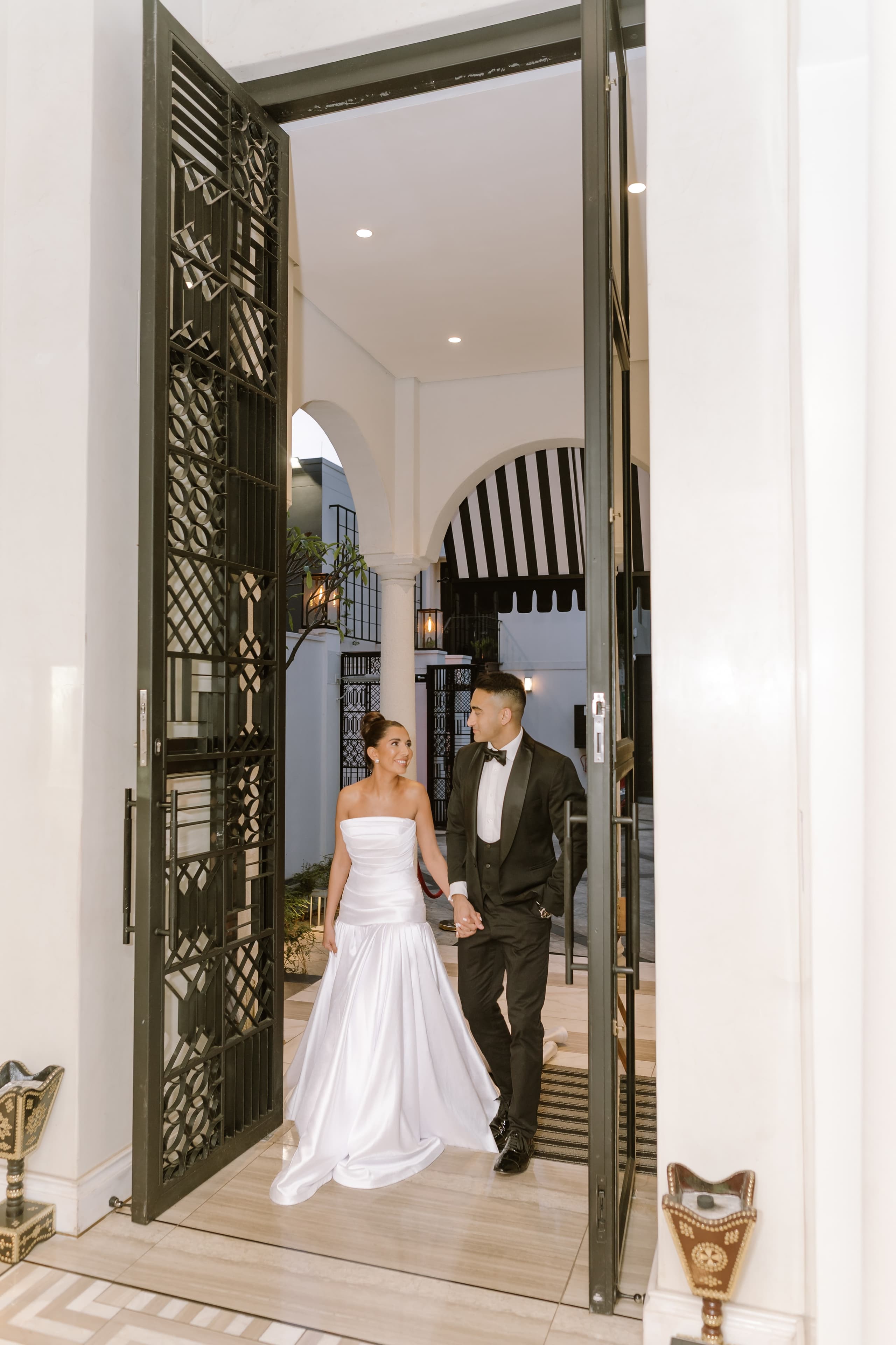 A bride and groom walk hand in hand through a grand doorway at sunset