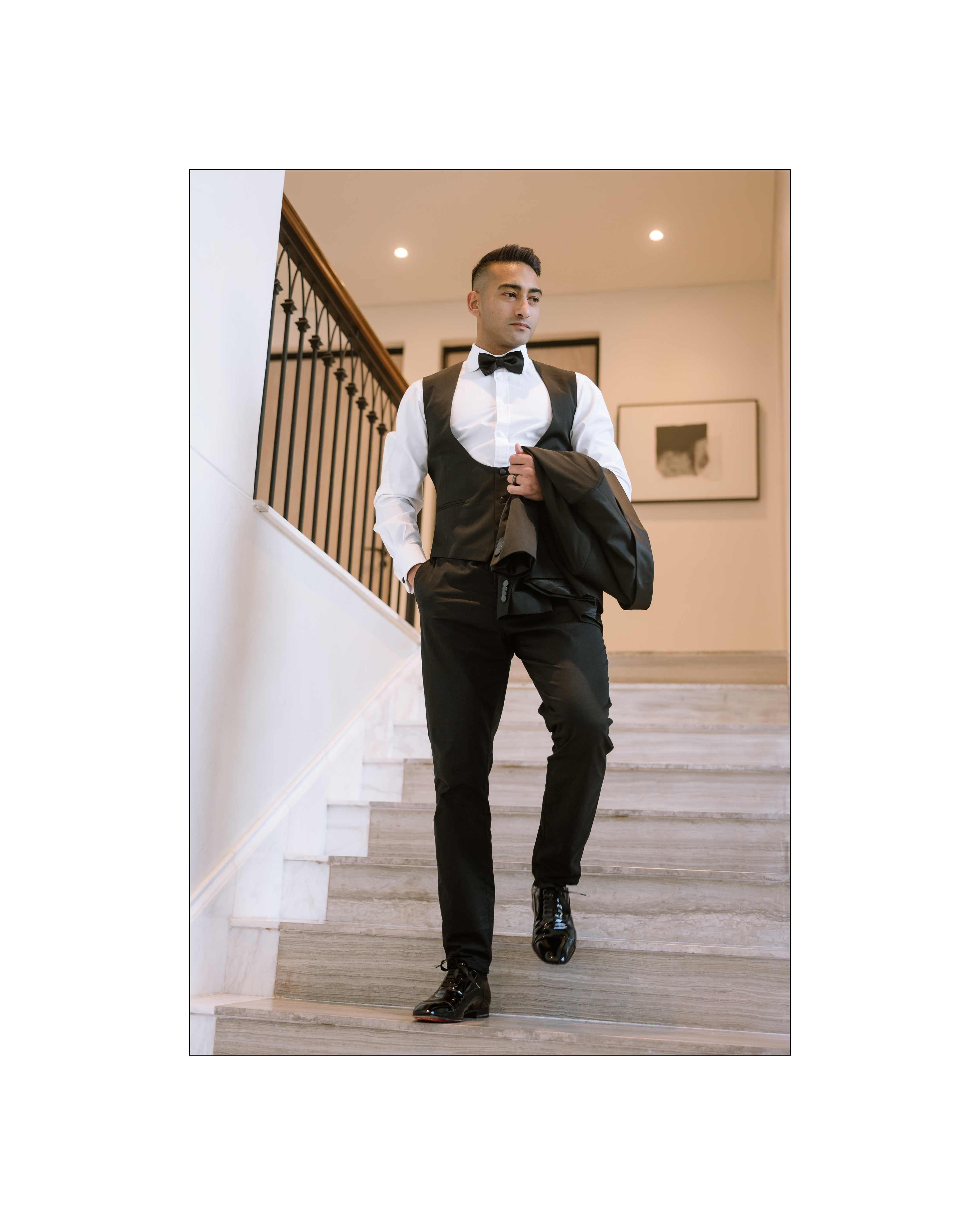A stylish groom in a black tuxedo vest and bowtie walks down a marble staircase