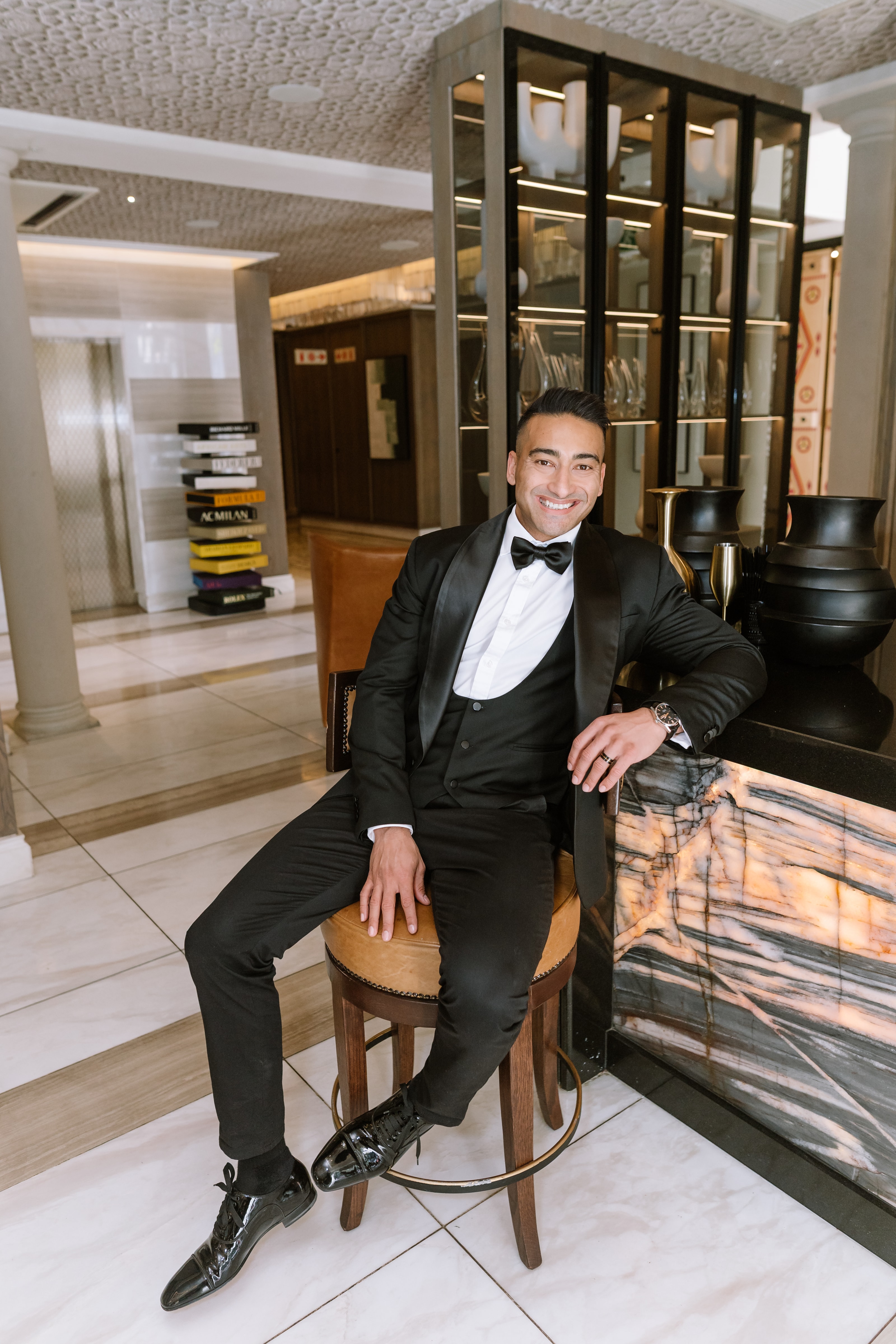 A smiling groom in a black tuxedo sits on a stool at a luxury bar