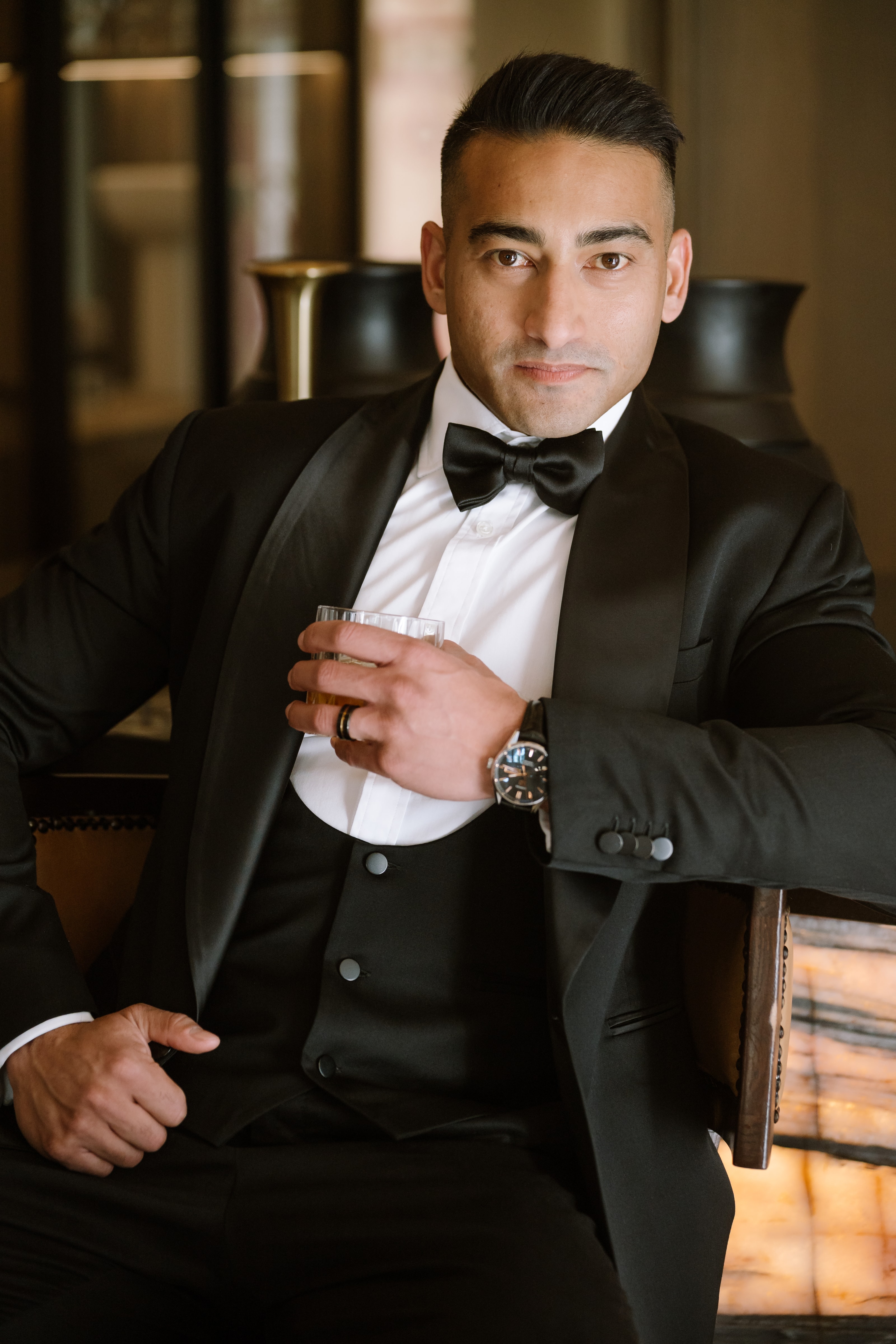 A dapper groom in a black tuxedo and bow tie holds a glass of whiskey